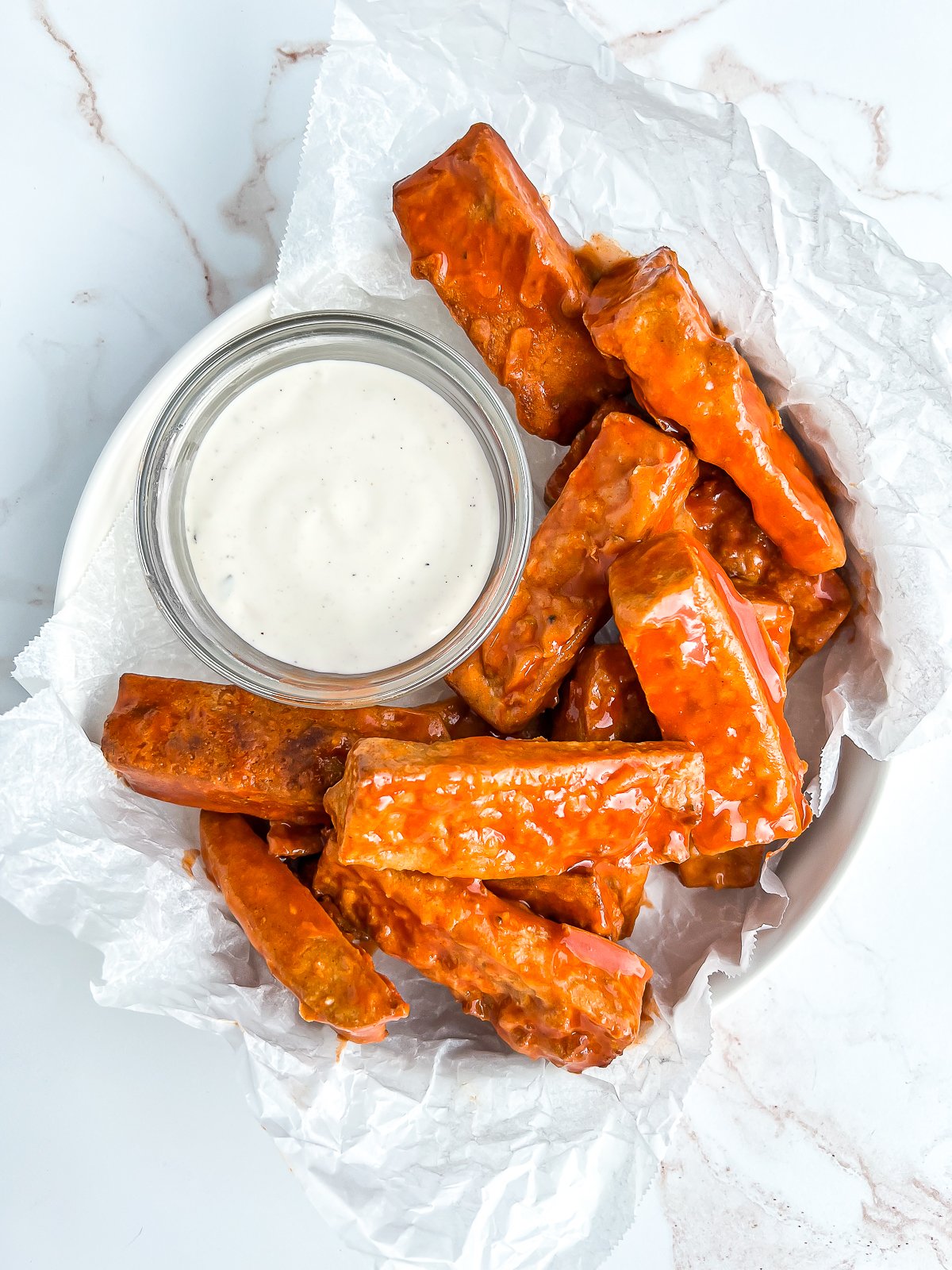 plate of tofu wing glistening from buffalo sauce, with a cup of ranch for dipping and crinkled parchment paper on a marble table