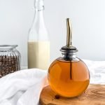 cinnamon dolce syrup in a glass container with a pour spout, with glass of milk and glass container of coffee beans behind it
