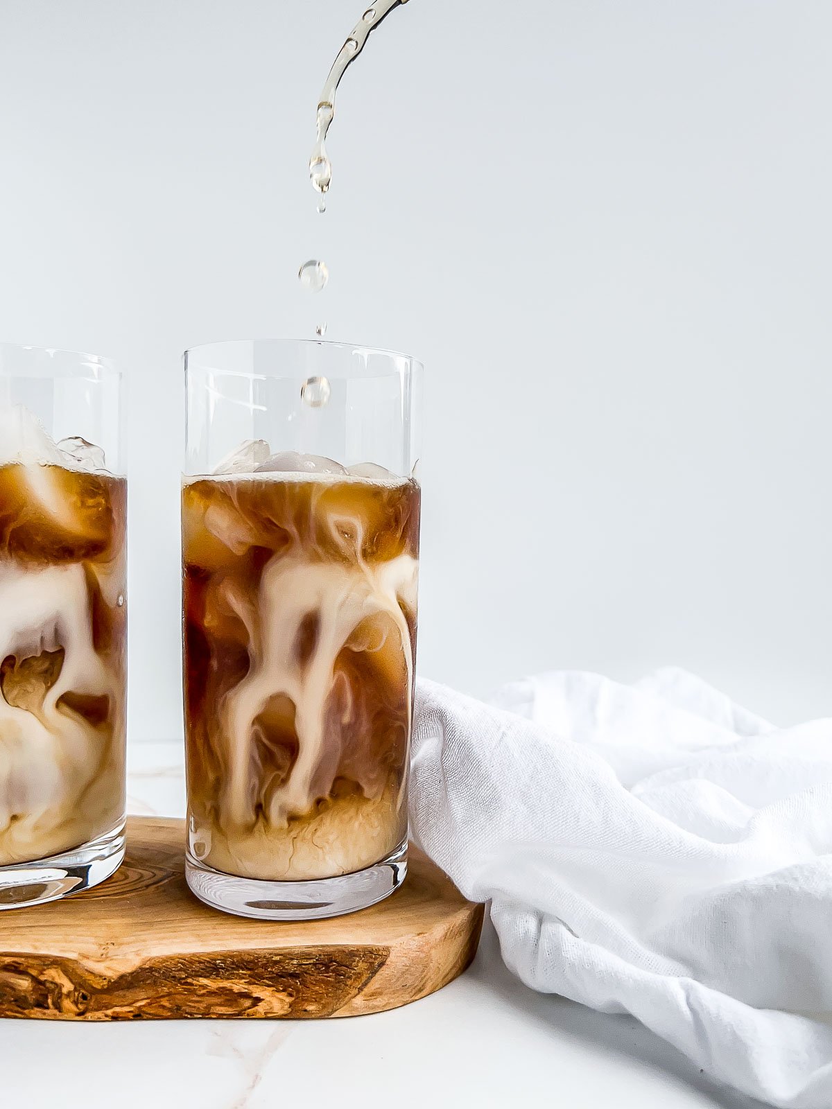 two glasses with iced coffee and swirls of milk, with syrup pouring into one glass