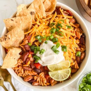 Bowl of chili mac topped with chips, sour cream, green onions, and limes.