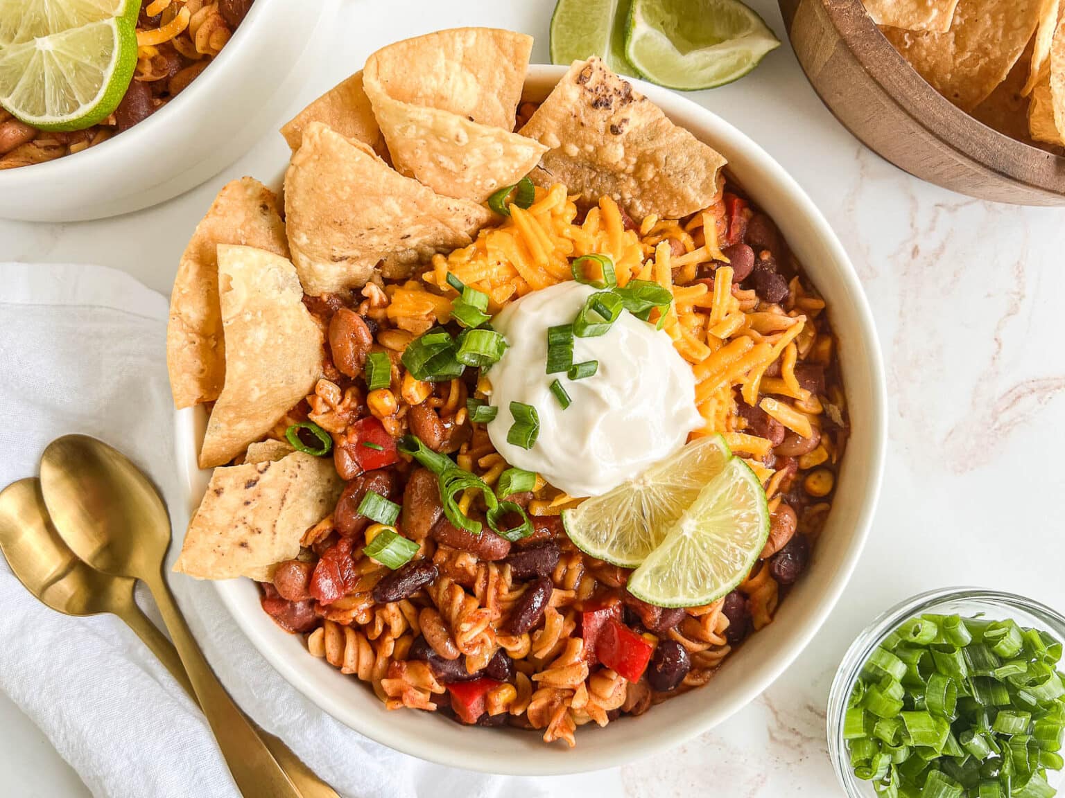 A bowl of chili pasta topped with shredded cheese, sour cream, green onions, and lime wedges. Tortilla chips are arranged on one side. A side of chopped green onions, lime wedges, a white napkin, and gold spoons are visible around the bowl.