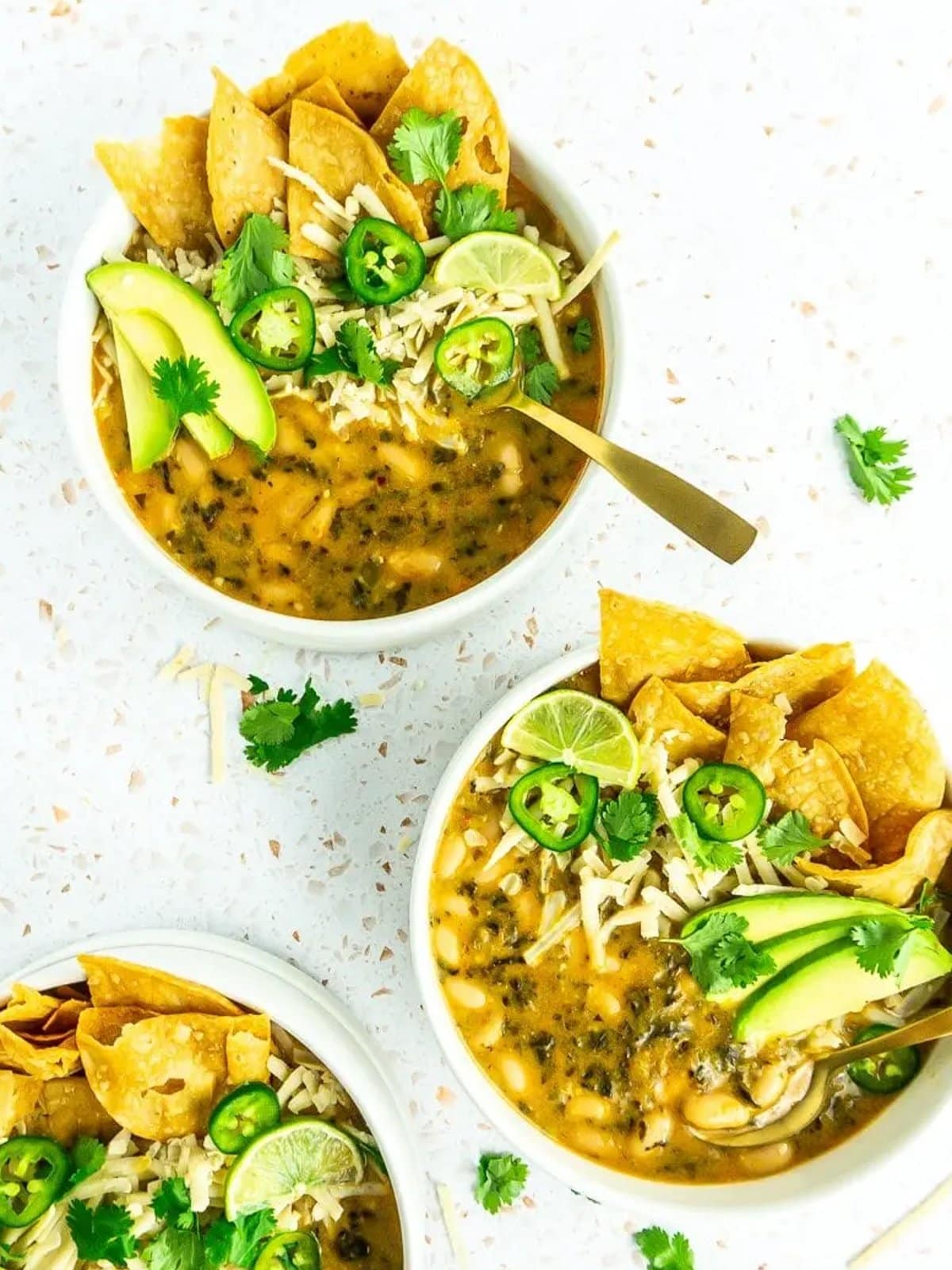 three bowls of white bean chili topped with cheese, jalapeños, chips, avocado, and fresh cilantro,