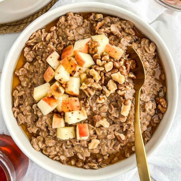 Very close-up image of a bowl of cinnamon spice oatmeal topped with chopped apples, walnuts, and maple syrup with a gold spoon tucked in
