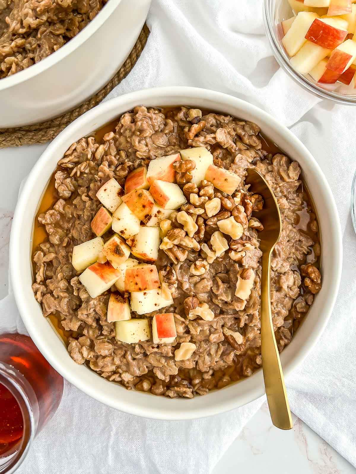 Bowl of cinnamon spice oatmeal topped with chopped apples, walnuts, and maple syrup with a gold spoon tucked in over a tea towel