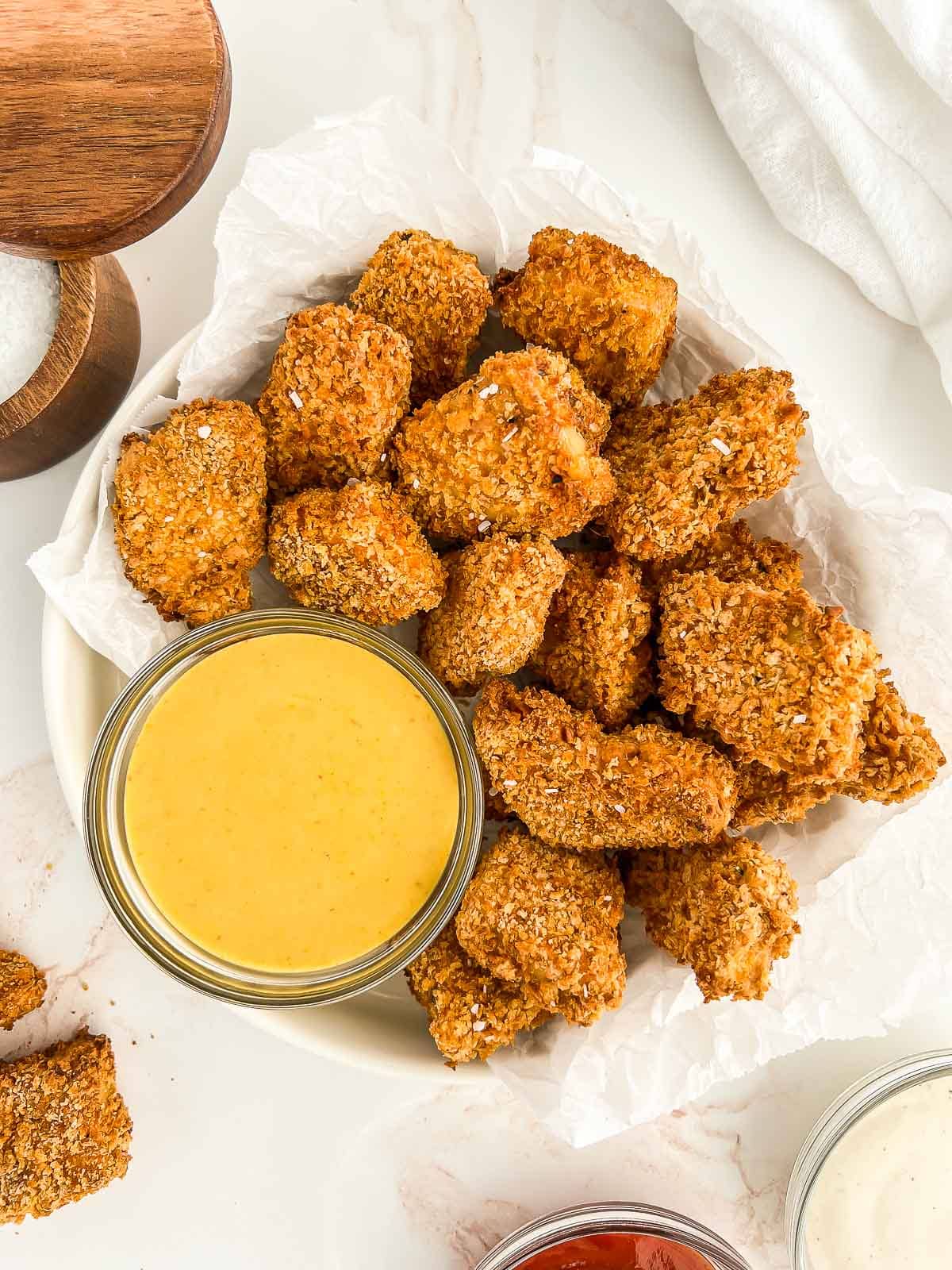 Bowl of tofu nuggets on parchment paper and dipping sauce, with other sauces and salt.