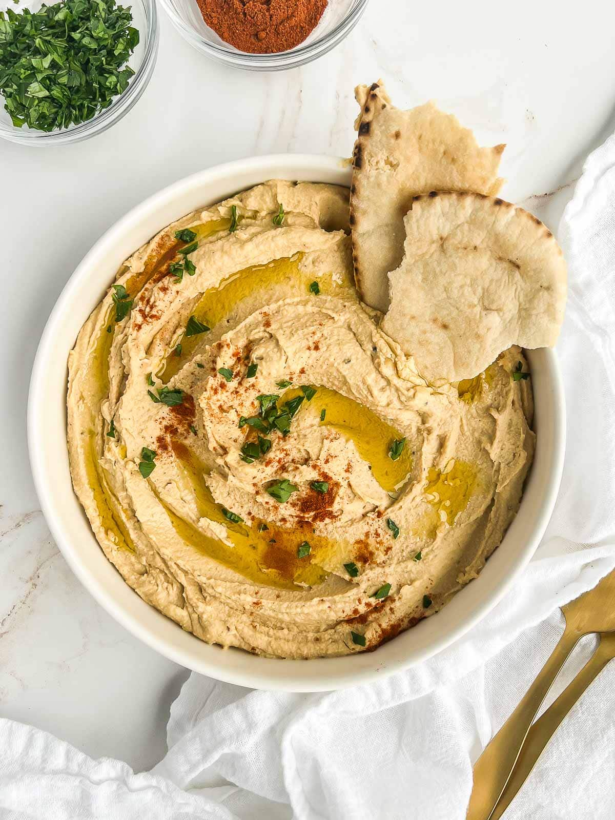Bowl of hummus topped with paprika, parsley, and olive oil with a couple pieces of pita tucked in.