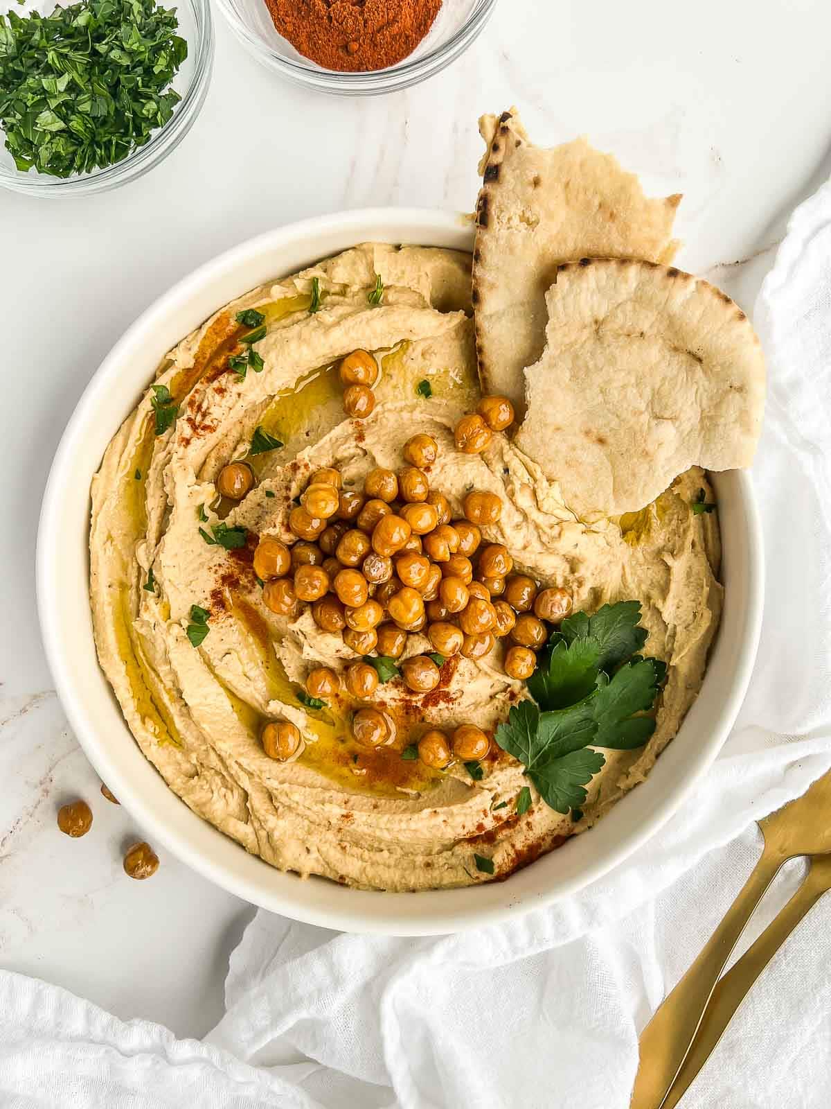 Bowl of hummus topped with crispy chickpeas, paprika, parsley, and olive oil with a couple pieces of pita tucked in.