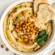 Bowl of hummus topped with crispy chickpeas, paprika, parsley, and olive oil with a couple pieces of pita tucked in.