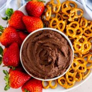 A cup of creamy chocolate hummus surrounded by pretzels and fresh strawberries.