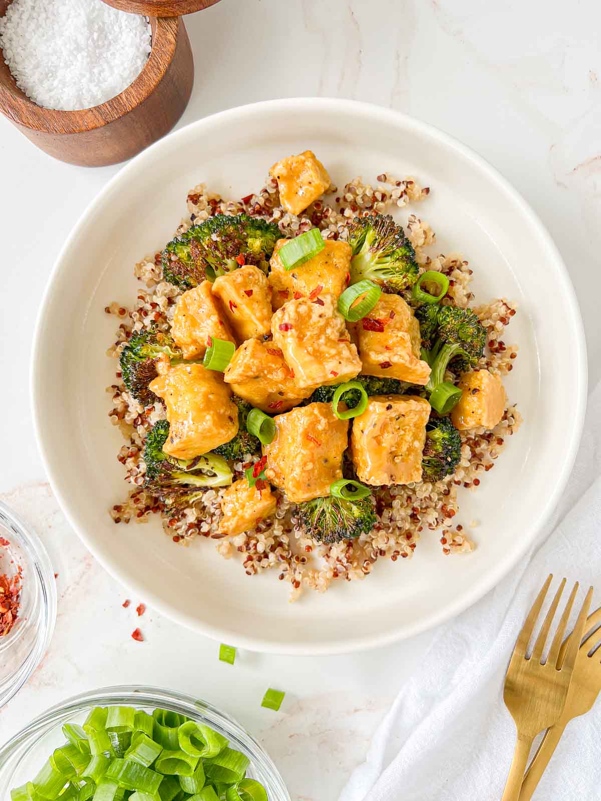 Plate of quinoa and roasted broccoli topped with vegan honey mustard tofu and garnished with green onions and red pepper flakes with salt well and gold forks nearby.