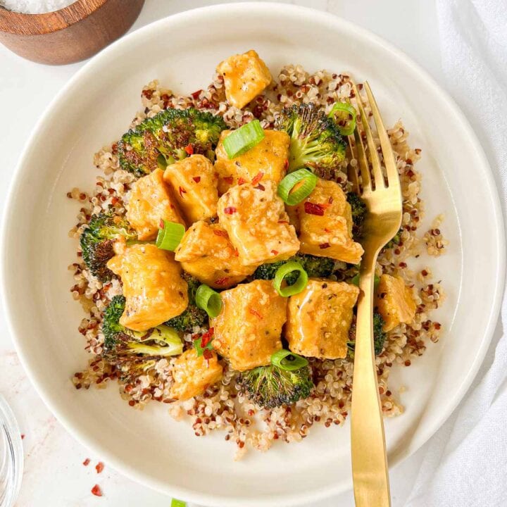 Plate of quinoa and roasted broccoli topped with vegan honey mustard tofu and garnished with green onions and red pepper flakes and a fork tucked in.
