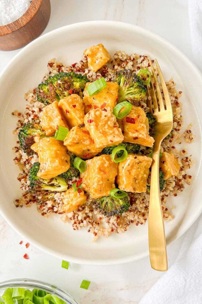 Plate of quinoa and roasted broccoli topped with vegan honey mustard tofu and garnished with green onions and red pepper flakes.