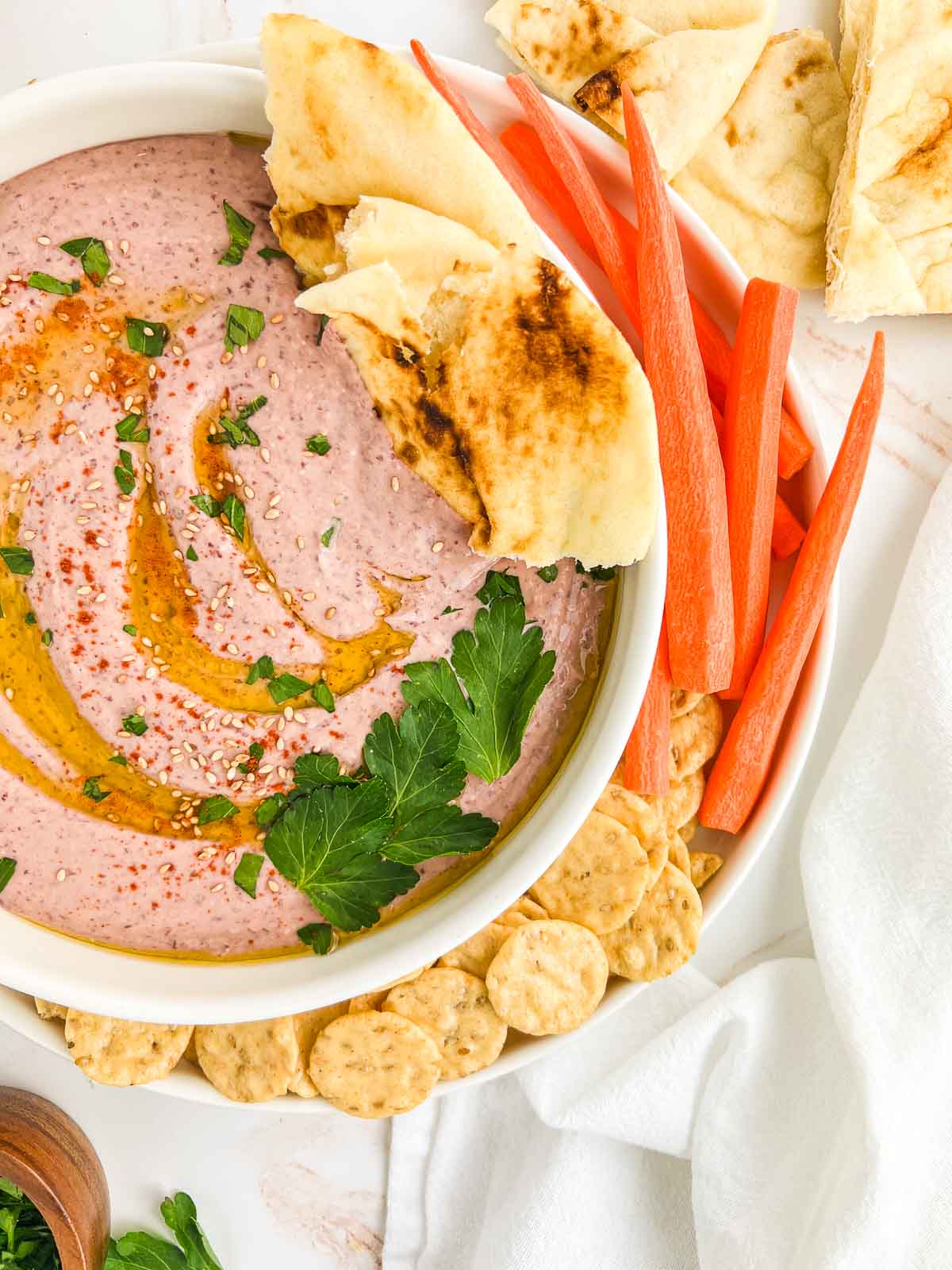 Bowl of kidney bean hummus garnished with oil, sesame seeds, and parsley, with carrots, crackers, and pita, with a white napkin.