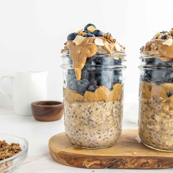 Two jars of overnight oats topped with peanut butter, blueberries, maple syrup, toasted coconut, and granola.